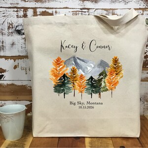 May include: A beige canvas tote bag featuring a watercolor design of mountains and trees in shades of orange, green, and gray. The text "Kacey & Connor" and "Big Sky, Montana 10.15.2026" are printed on the bag.