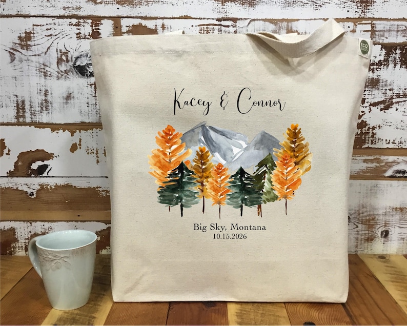 May include: A beige canvas tote bag featuring a watercolor design of mountains and trees in shades of orange, green, and gray. The text "Kacey & Connor" and "Big Sky, Montana 10.15.2026" are printed on the bag.