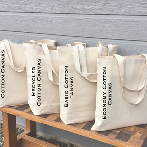 May include: Four different types of tote bags made from cotton canvas. The bags are lined up on a wooden bench. The bags are labeled "Large Recycled Cotton Canvas", "Recycled Cotton Canvas", "Basic Cotton Canvas", and "Economy Cotton Canvas".