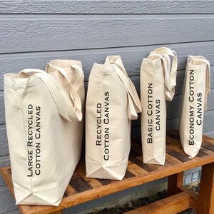 May include: Four different types of tote bags lined up on a wooden surface. From left to right, the bags are labeled "Large Recycled Cotton Canvas", "Recycled Cotton Canvas", "Basic Cotton Canvas", and "Economy Cotton Canvas".
