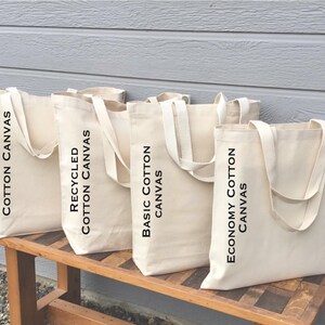 May include: Four beige canvas tote bags with white handles are displayed on a wooden bench. The bags are labeled with black text: "Large Recycled Cotton Canvas", "Recycled Cotton Canvas", "Basic Cotton Canvas", and "Economy Cotton Canvas". The background is a light gray wall.