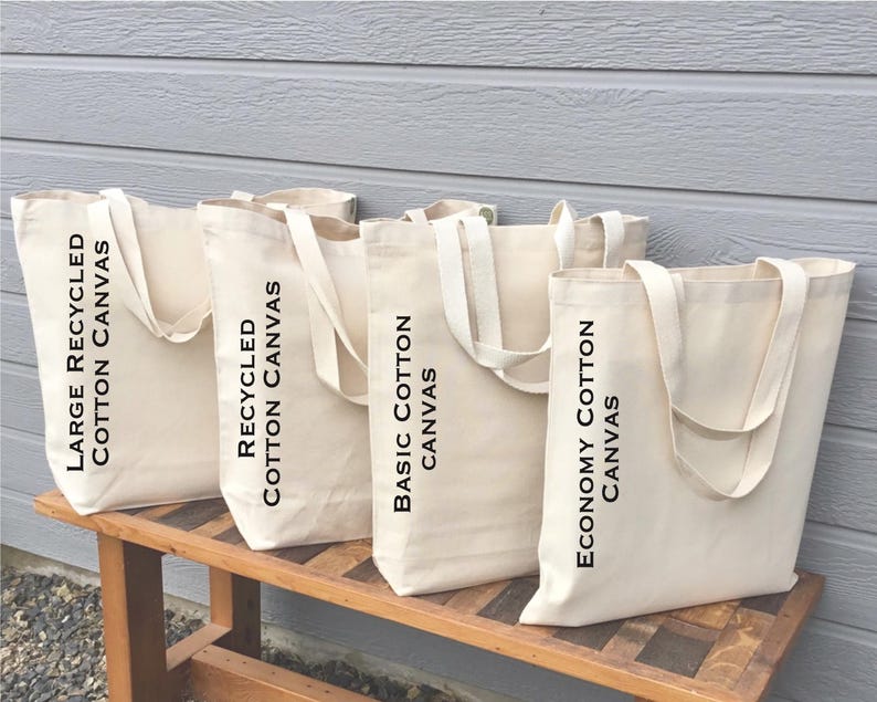 May include: Four beige canvas tote bags with white handles are displayed on a wooden bench. The bags are labeled with black text: "Large Recycled Cotton Canvas", "Recycled Cotton Canvas", "Basic Cotton Canvas", and "Economy Cotton Canvas". The background is a light gray wall.