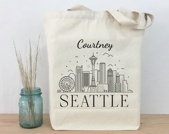 May include: A white canvas tote bag with black text and a line drawing of the Seattle skyline, including the Space Needle. The text on the bag reads "Courtney Seattle".