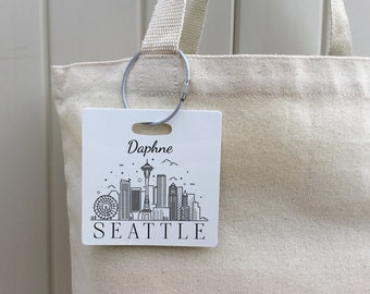 Könnte beinhalten: Weiße Canvas-Tragetasche mit einem grauen Metallring und einem weißen Anhänger. Der Anhänger zeigt eine Schwarzweiß-Illustration der Skyline von Seattle mit dem Space Needle und dem Text "Daphne Seattle".