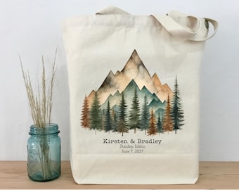 Könnte beinhalten: Cremefarbene Tragetasche mit einem Aquarell-Berg- und Waldmotiv in Braun-, Grün- und Orangetönen. Die Tasche hat Henkel und die Namen "Kirsten & Bradley" sowie das Datum "7. Juni 2027" sind aufgedruckt.