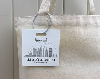 Könnte beinhalten: Weiße Canvas-Tragetasche mit einem grauen Metallring und einem weißen Anhänger. Der Anhänger zeigt eine Schwarzweiß-Illustration der Skyline von San Francisco und den Text "Hannah San Francisco Kalifornien".