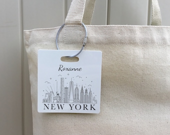 Könnte beinhalten: Eine weiße Tragetasche mit einem schwarz-weißen New Yorker Skyline-Gepäckanhänger. Der Anhänger trägt den Text "Roxanne" und "NEW YORK" in schwarzer Schrift.