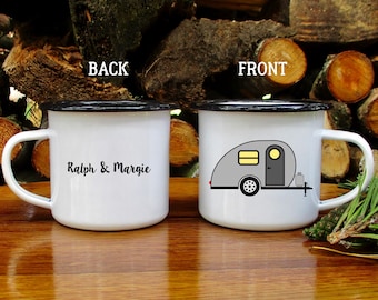 Könnte beinhalten: Weißer Emaille-Campingbecher mit schwarzem Rand und Henkel. Die Vorderseite zeigt eine graue Wohnwagen-Illustration mit gelbem Fenster und Tür. Auf der Rückseite des Bechers steht der Text "Ralph & Margie" in schwarzer Schrift.