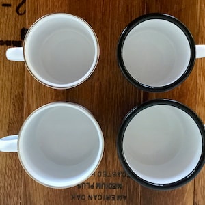 May include: Four white enamel mugs with contrasting black and gold rims, each with a white handle. The mugs are arranged on a wooden surface, offering a top-down view of the interiors.