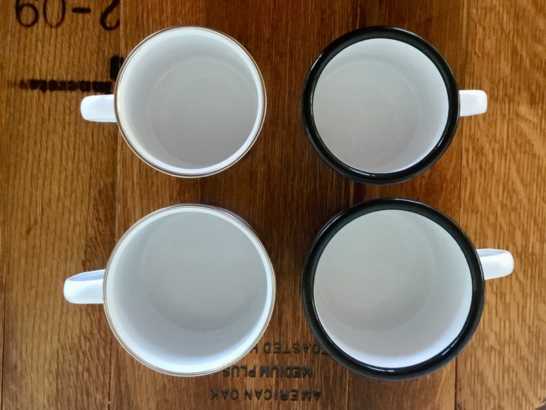 May include: Four white enamel mugs with contrasting black and gold rims, each with a white handle. The mugs are arranged on a wooden surface, offering a top-down view of the interiors.