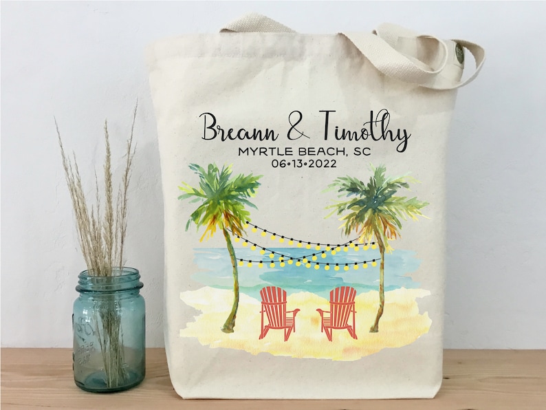 May include: A white canvas tote bag with a beach scene design. The bag features two palm trees, a beach with two red chairs, and string lights. The text "Breann & Timothy" is printed on the bag, along with "Myrtle Beach, SC" and "06-13-2022".