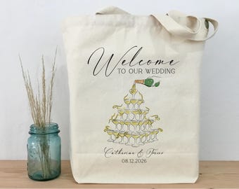 Könnte beinhalten: Cremefarbene Tragetasche mit dem Schriftzug "Welcome to our wedding" in eleganter Schrift. Darunter eine Illustration eines Champagnerturms, mit den Namen "Catherine & Trevor" und dem Datum "08.12.2026". Ein Hochzeitsaccessoire.