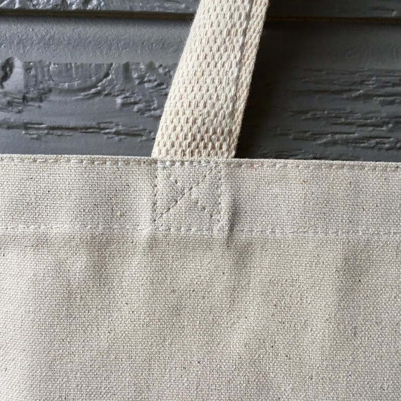 May include: Close-up of a natural canvas tote bag with a cream-colored handle. The bag is made of a sturdy, textured fabric, with reinforced stitching at the handle attachment points. The handle is a woven, cream-colored material.