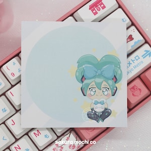 May include: A light blue sticker with a cartoon illustration of a girl with green hair wearing a blue bow and holding a white plushie. The sticker is on a white background with a pink keyboard in the background.
