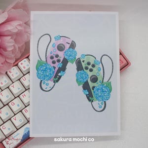 May include: A white background with two Nintendo Switch controllers, one pink and one green, with blue hydrangea flowers around them. The text "sakura mochi co" is at the bottom of the image.