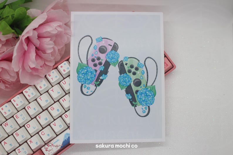 May include: A white background with two Nintendo Switch controllers, one pink and one green, with blue hydrangea flowers around them. The text "sakura mochi co" is at the bottom of the image.