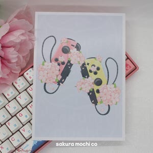 May include: Two Nintendo Switch controllers decorated with pink and yellow flowers. The controllers are in a heart shape with the pink controller on the left and the yellow controller on the right. The background is a light blue color.