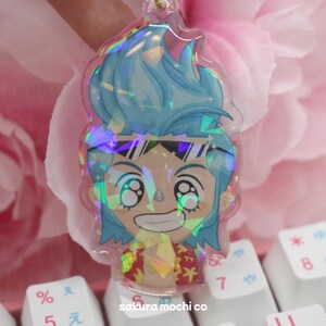 May include: A holographic charm featuring a cartoon character with blue hair, wearing sunglasses and a patterned shirt. The charm is on a white background with a keyboard in the background.