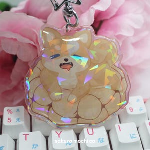 May include: A clear acrylic keychain featuring a cartoon Shiba Inu dog with a holographic effect. The dog is light brown and cream-colored, with its tongue sticking out. The keychain is on a keyboard with Japanese characters.