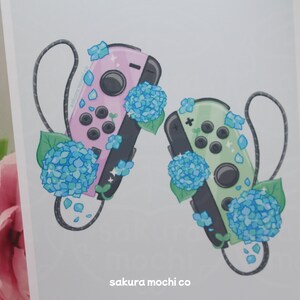 May include: Two Nintendo Switch controllers, one pink and one green, with blue hydrangea flowers. The controllers are decorated with glitter and the flowers have a sparkly finish. The text "sakura mochi co" is at the bottom of the image.