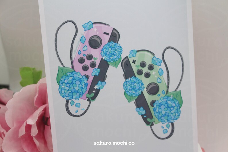May include: Two Nintendo Switch controllers, one pink and one green, with blue hydrangea flowers. The controllers are decorated with glitter and the flowers have a sparkly finish. The text "sakura mochi co" is at the bottom of the image.