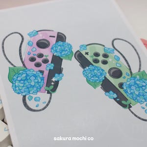 May include: Two Nintendo Switch controllers, one pink and one green, are surrounded by blue hydrangea flowers. The controllers are decorated with white and blue glitter. The image includes the text "sakura mochi co."