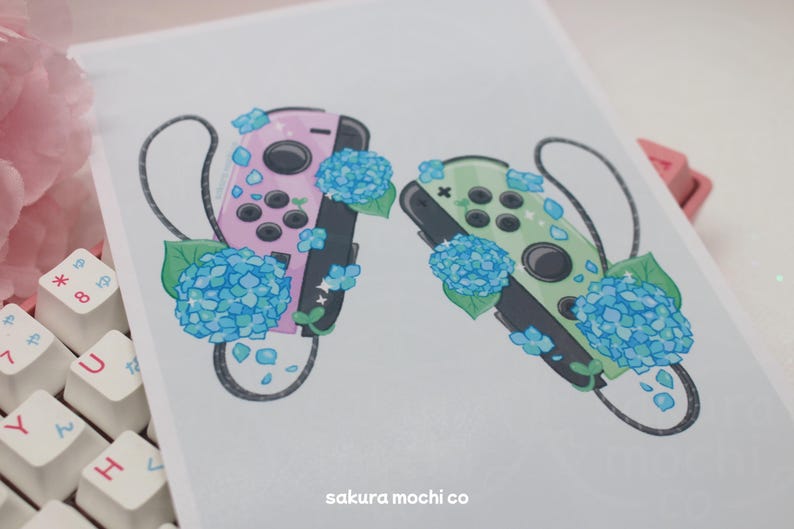 May include: Two Nintendo Switch controllers, one pink and one green, are surrounded by blue hydrangea flowers. The controllers are decorated with white and blue glitter. The image includes the text "sakura mochi co."