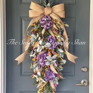 May include: A decorative teardrop-shaped floral arrangement with purple hydrangeas, yellow accents, and green foliage, accented by a large burlap bow. The arrangement hangs on a blue door. Text on the image reads "The Swag Boutique by Shan."