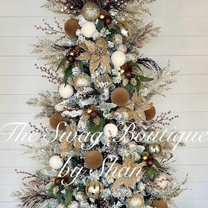 May include: A decorated Christmas tree with white, brown, and gold ornaments. The tree features white and brown spherical ornaments, gold floral accents, and frosted branches. The text "The Swag Boutique by Shan" is visible.