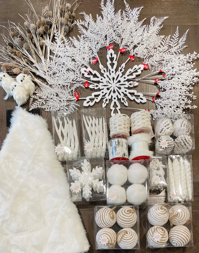 May include: White winter-themed decorations including a snowflake, a star, and a variety of ornaments. The decorations are arranged on a wooden surface and include white branches, white feathers, and white ribbon.