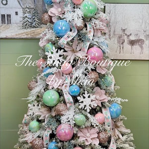 May include: A decorated Christmas tree with pastel ornaments, ribbons, and floral accents. The tree features blue, pink, and green baubles, with a woven basket base. The text "The Frugal Boutique by Shan" is visible.