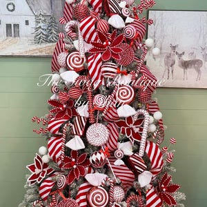 May include: A Christmas tree decorated with red and white candy-themed ornaments, including striped lollipops, candy canes, wrapped hard candies, and red poinsettia flowers. The tree features white-tipped branches and a text overlay reading The Swag Boutique by Dian.
