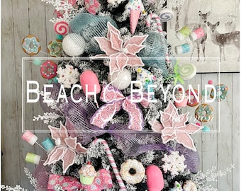 Puede incluir: Un árbol de Navidad rosa y azul decorado con bastones de caramelo, donas, cupcakes y otras golosinas dulces. El árbol está coronado con una gran piruleta rosa. El árbol está decorado con el texto "Beach & Beyond".