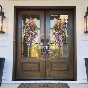 May include: Two wooden doors with glass panels are adorned with floral teardrop swags. Each swag features purple, white, and green flowers, accented with a tan bow. The text "THE SWAG BOUTIQUE by Shan" is visible on the door.