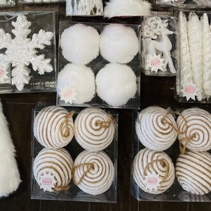 May include: Assortment of white and brown Christmas ornaments. The ornaments are in various shapes, including snowflakes, balls, and swirls. The ornaments are displayed in clear plastic containers.