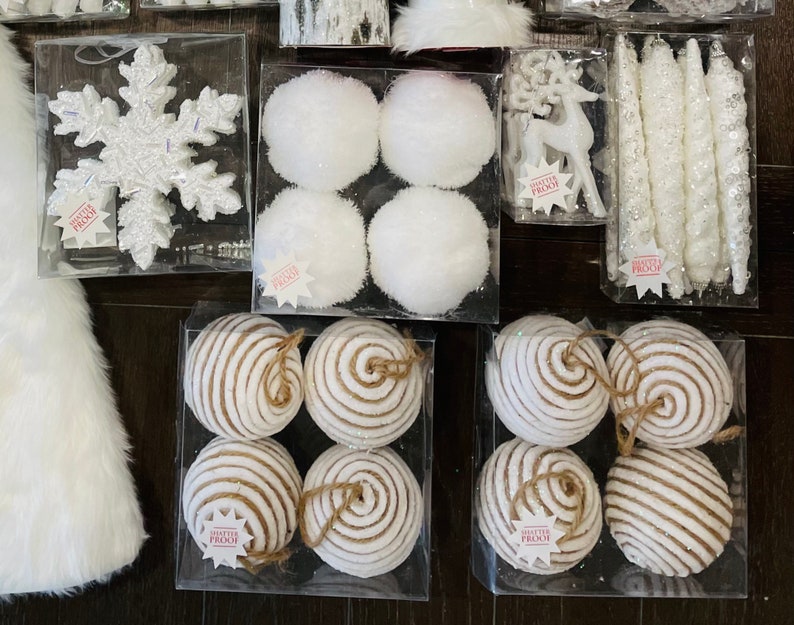 May include: Assortment of white and brown Christmas ornaments. The ornaments are in various shapes, including snowflakes, balls, and swirls. The ornaments are displayed in clear plastic containers.
