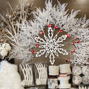 May include: A collection of white winter-themed decorations, including a snowflake, branches, and a faux fur rug. The decorations are arranged on a dark wood surface.