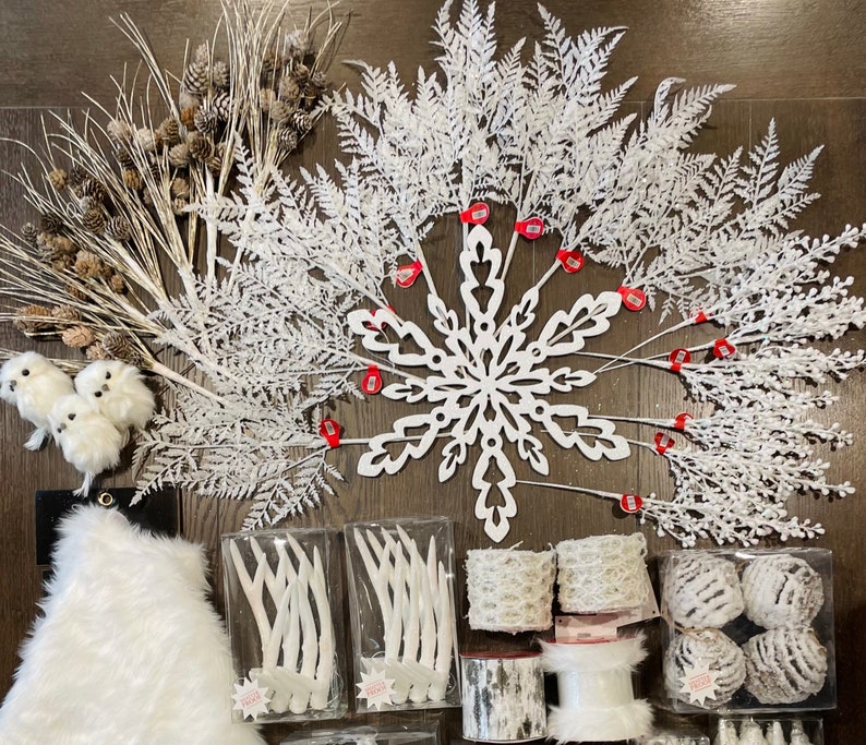May include: A collection of white winter-themed decorations, including a snowflake, branches, and a faux fur rug. The decorations are arranged on a dark wood surface.
