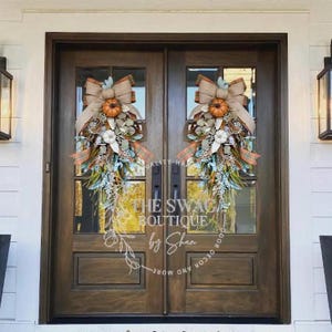 May include: Two brown wooden doors with glass panels, each adorned with a fall-themed wreath. The wreaths feature pumpkins, bows, and foliage in orange, brown, and green hues. The text "THE SWAG BOUTIQUE by Shan" is visible on the doors.