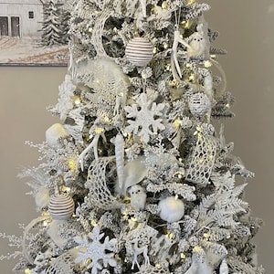 May include: A white flocked Christmas tree decorated with white and silver ornaments, including snowflakes, deer, owls, and a white mesh ribbon.
