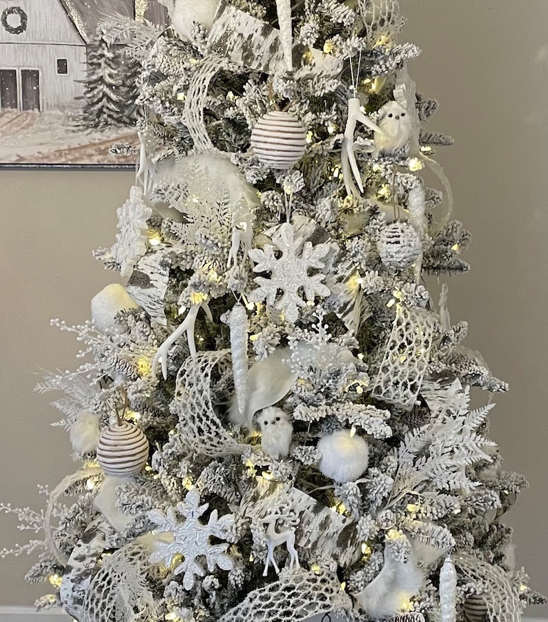 May include: A white flocked Christmas tree decorated with white and silver ornaments, including snowflakes, deer, owls, and a white mesh ribbon.