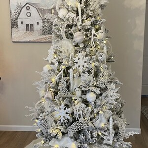 May include: A white flocked Christmas tree decorated with white and brown ornaments, white birds, and a white snowflake tree topper. The tree is standing on a white faux fur tree skirt.