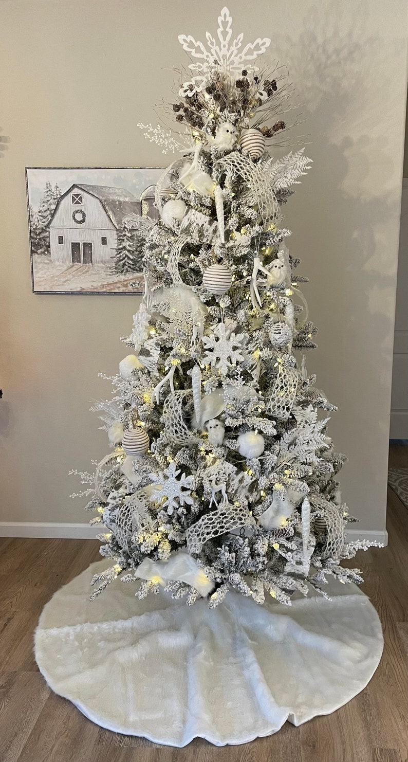 May include: A white flocked Christmas tree decorated with white and brown ornaments, white birds, and a white snowflake tree topper. The tree is standing on a white faux fur tree skirt.