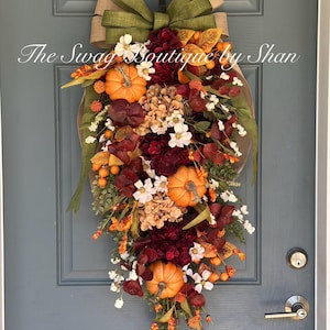 May include: A teardrop-shaped autumn door swag featuring three orange pumpkins, deep burgundy and tan hydrangea blooms, white flowers, and green foliage. A large olive green and tan burlap bow sits at the top. Text reads The Swag Boutique by Shan.