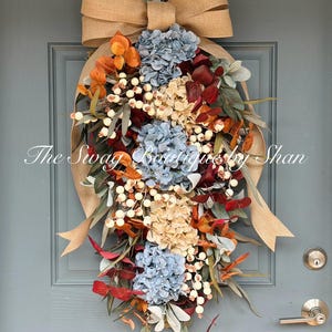 May include: A fall-themed wreath with blue, beige, and red flowers, white berries, and green leaves. The wreath is adorned with a large burlap bow and is hanging on a blue door. The text "The Swag Boutiques by Shan" is visible.
