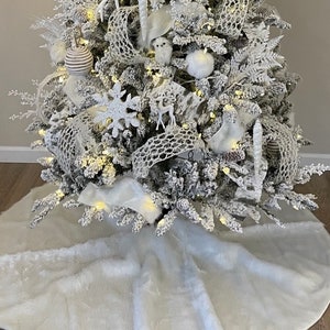 May include: A white faux fur Christmas tree skirt with a white flocked Christmas tree decorated with white and silver ornaments, lights, and ribbon.