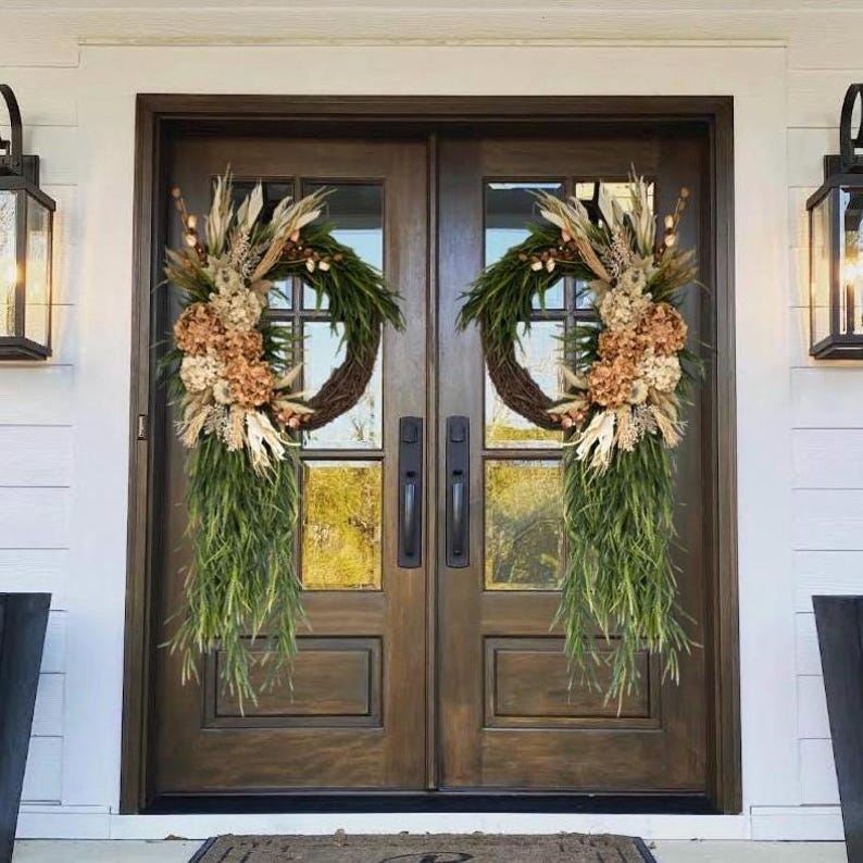 May include: Two brown double doors with glass panels and black door handles. Each door is decorated with a green and beige floral wreath with dried flowers and long green leaves hanging down.