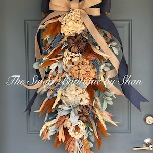 May include: A teardrop-shaped floral swag featuring dried hydrangea blossoms, rust-colored pampas grass, and sage green foliage. The arrangement is accented with a large layered bow made of navy blue and tan burlap ribbons, with the text The Swag Boutique by Shan.