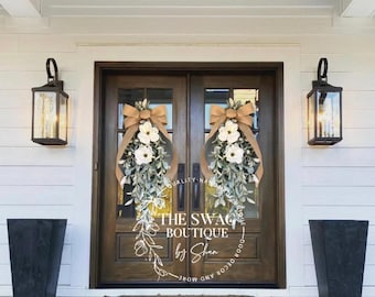 May include: A wooden front door with two wreaths, each with white flowers, greenery, and a tan bow. The door has the text "THE SWAG BOUTIQUE by Shan". Black lanterns flank the door, and black planters sit on either side.