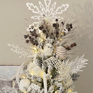 May include: A white flocked Christmas tree decorated with white and brown ornaments, a white snowflake tree topper, and white owls. The tree is lit with white fairy lights.